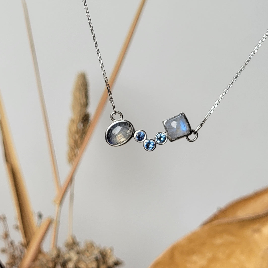 Oval moonstone, three Montana sapphires, and square moonstone pendant in fron of white background with yellow folliage