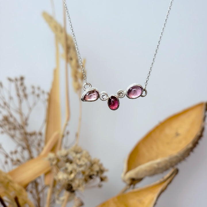 Magenta and baby pink tourmalines with white sapphires making a cluster necklace that is in front of a white backgrount with yellow folliage